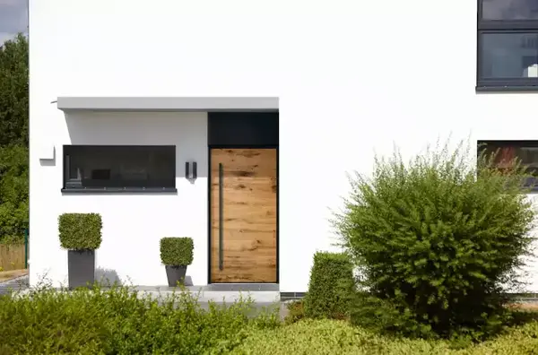 Moderne Haustür aus Holz in weißer Fassade, umgeben von gepflegten Pflanzen und Sträuchern.