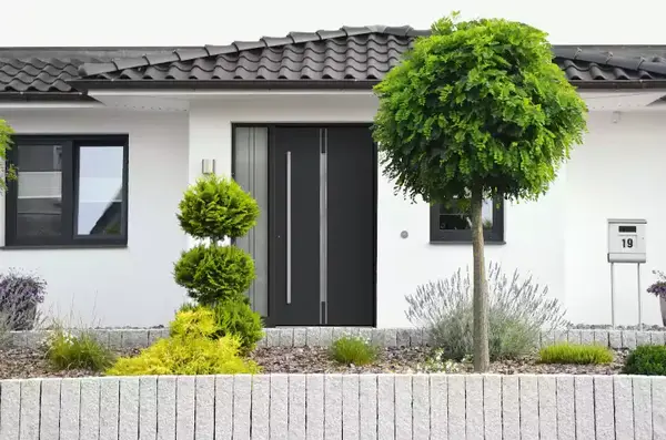 Moderner Hauseingang mit dunkler Haustür, gepflegtem Vorgarten und dekorativen Bäumen.